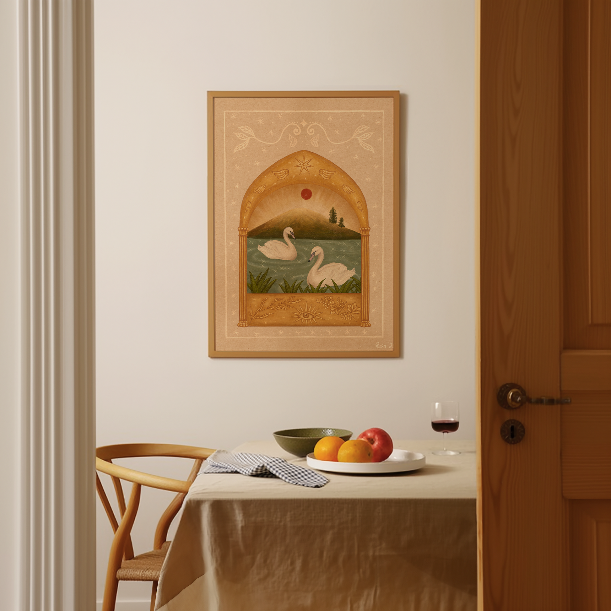 Framed fine art print of two white swans on a teal sparkly lake under a red moon and a golden arch. Frame on a white wall above a table and chair with food and wine.